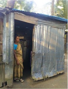 Woman in a sari leans against the door of her house. It is a one story metal building