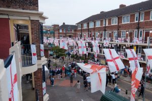 London estate with many England flags strung between buildings. On the street are a crowd of England fans watching the Euro 2020 final